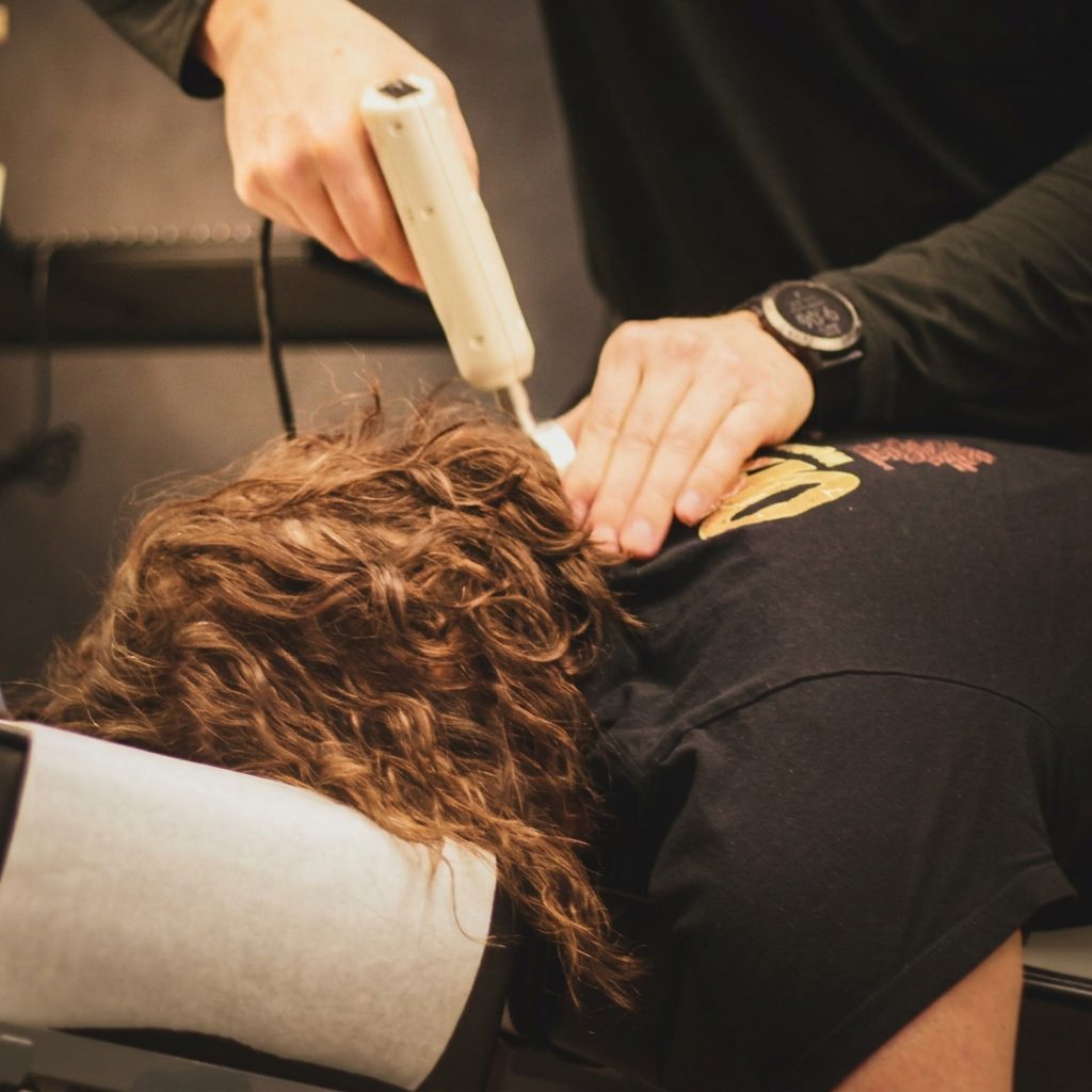 Practitioner using an electronic impulse adjusting gun on a patient's upper back and neck.
